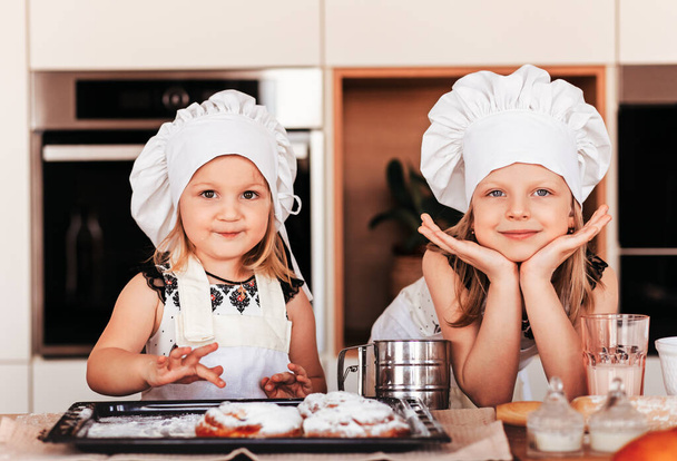 Δύο μικρά κορίτσια με άσπρα καπέλα σεφ διασκεδάζουν στην κουζίνα μαζί. - Φωτογραφία, εικόνα