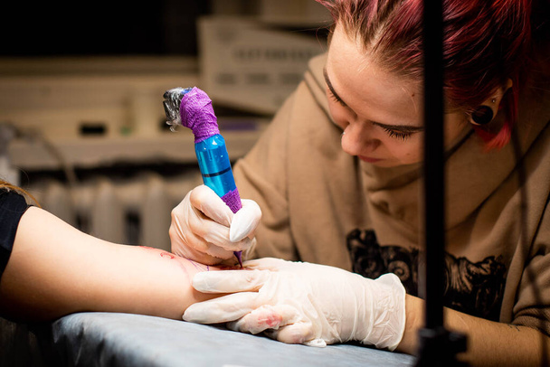 Mester tetoválás rajzolja a piros festék az ügyfelek tetoválás. Tetováló művész kezében egy rózsaszín tetováló gép fekete steril kesztyű és dolgozik a professzionális kék szőnyeg. - Fotó, kép