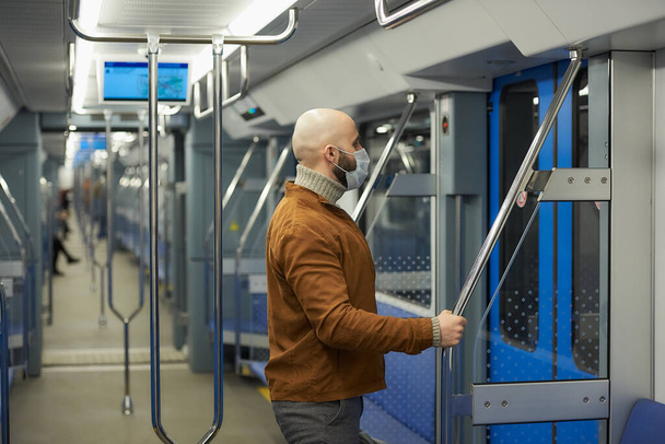 Egy szakállas férfi orvosi maszkban, hogy elkerülje a koronavírus terjedését, elhagyja a metrókocsit a korláttal a kezében. Egy maszkos kopasz fickó tartja a távolságot a vonaton.. - Fotó, kép