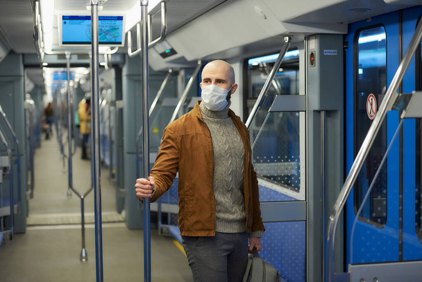 Egy szakállas férfi orvosi maszkban, hogy elkerülje a koronavírus terjedését, egy metrókocsiban ül és tartja a korlátot. Egy kopasz fickó sebészeti maszkban távolságot tart a vonaton.. - Fotó, kép