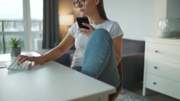 Nenuceně oblečená žena se sluchátky pracující s počítačem a smartphonem doma v útulném prostředí. Vzdálená práce mimo kancelář. - Záběry, video
