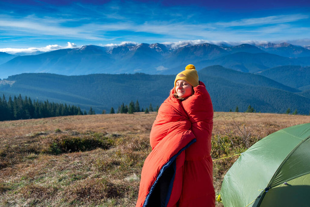 Młoda dziewczyna stoi ze śpiworem w pobliżu namiotu z górami na tle - Zdjęcie, obraz