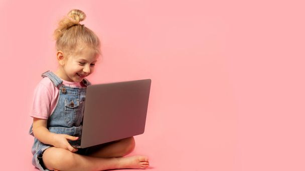 Usmívající se holčička sedí s notebookem izolovaným na růžovém pozadí. Školní distanční vzdělávání doma během karanténní koncepce. nápis s mezerou pro text. - Fotografie, Obrázek