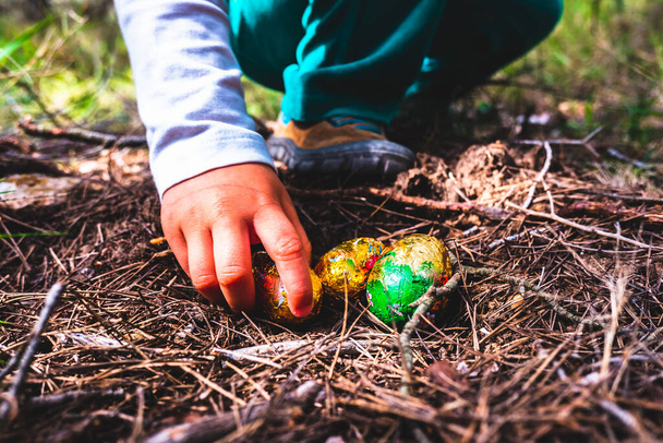 La mano de una chica recoge un huevo de pascua de chocolate encontrado en un bosque. - Foto, Imagen