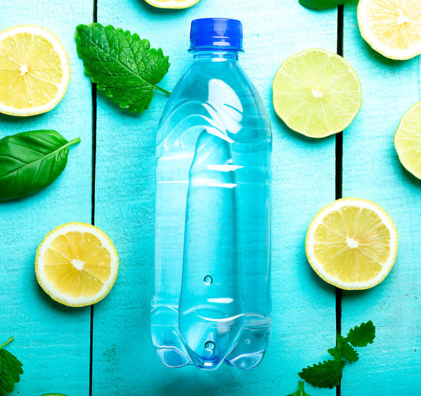 Бутылка чистой воды, лежащая на деревянной бирюзовой поверхности с лимонами, мятой и льдом, крупным планом - Фото, изображение
