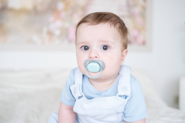 close up portrait baby boy 6 months old with nipple in blu clothes and sitting on white bed at home - Foto, Imagen