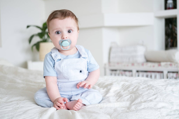 baby jongen 6 maanden oud met tepel in blu kleding en zitten op wit bed thuis - Foto, afbeelding