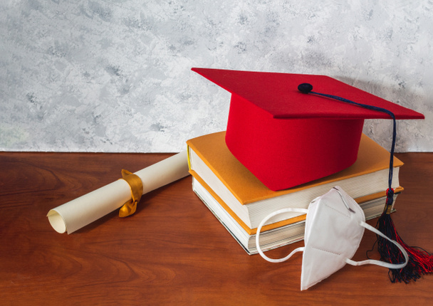 Stilleven met een rode afstudeerhoed op enkele boeken naast een diploma en een masker op een houten bureau. Concept van afstuderen en sociale afstand - Foto, afbeelding