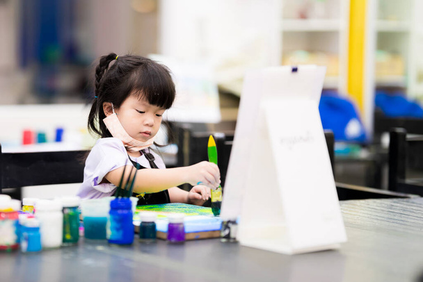 Cute 4 year old Asian girl doing crafts on canvas. Child learn art in class. Children use large paintbrush to gently dip watercolors onto work board. Kid take cloth mask under chin. Pupil enjoy hobby. - Photo, Image