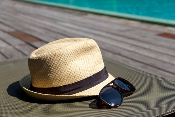 Κοντινό πλάνο ψάθινο καπέλο και γυαλιά ηλίου στο κρεβάτι δίπλα στην πισίνα. Διακοπές για τις καλοκαιρινές διακοπές έννοια. - Φωτογραφία, εικόνα