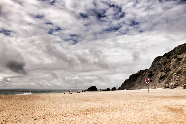シントラ、リスボン、ポルトガルのアドラガのビーチで勇敢な海、岩の形成と曇りのドラマの空 - 写真・画像