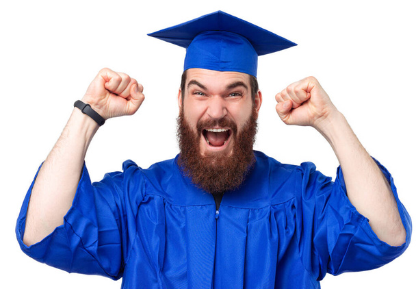Ενθουσιασμένος και φωνάζοντας φοιτητής με γενειάδα φορώντας εργένικο και καπέλο αποφοίτησης γιορτάζει. - Φωτογραφία, εικόνα