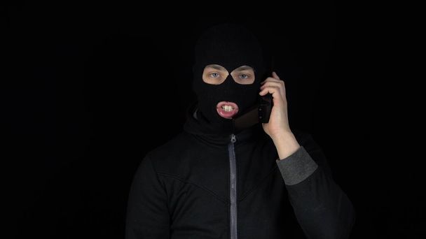 A man in a balaclava mask with a phone from the 90s. The bandit is talking on the phone. On a black background. - Фото, изображение