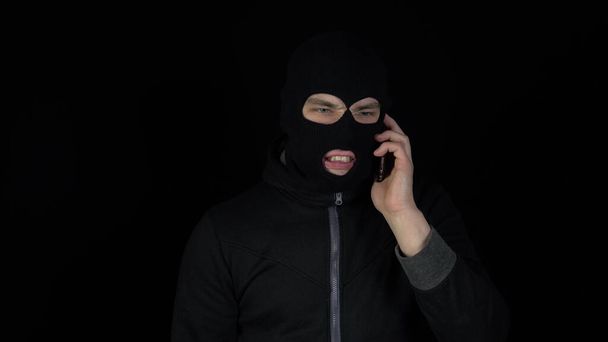 Um homem com uma máscara de balaclava está a falar ao telefone. Um bandido gritando ao telefone em um fundo preto. - Foto, Imagem