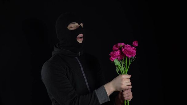 Un uomo con una maschera a passamontagna e 'in piedi con dei fiori. Bandit annusa un mazzo di fiori rosa su sfondo nero. - Foto, immagini