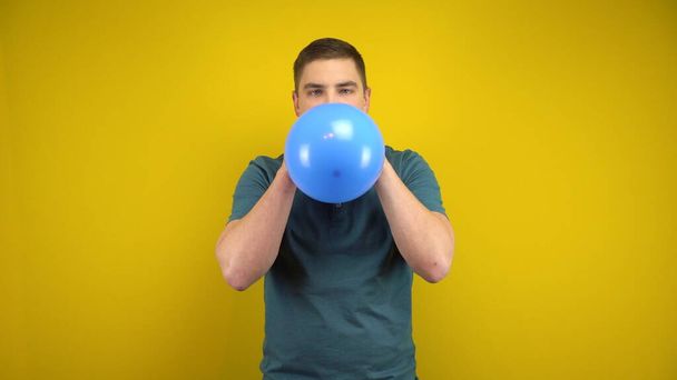 Молодой человек надувает синий шарик ртом на жёлтом фоне. Человек в зелёном поло. - Фото, изображение