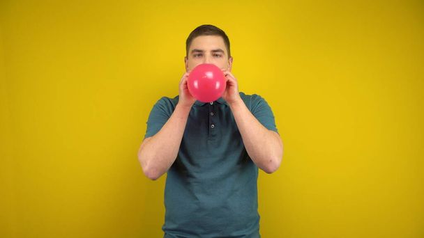 Um jovem infla um balão vermelho com a boca em um fundo amarelo. Homem num pólo verde. - Foto, Imagem