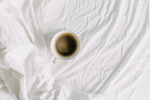 Вид сверху на чашку черного кофе на белой простыне. завтрак в кровать концепции. - Фото, изображение