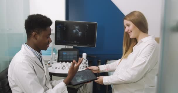 Lääkäri ja potilas ultraäänihuoneessa. Afrikkalainen mieslääkäri puhuu nuorelle raskaana olevalle naiselleen ultraäänitoimenpiteen jälkeen ja kertoo ultraäänikokeen tuloksista, jotka osoittavat skannauksen tabletilla - Materiaali, video