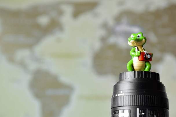 Декоративная фигурка крокодила с камерой, стоящего на объективе на фоне размытой карты мира с правой стороны рамки. - Фото, изображение