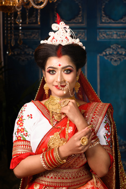 Impresionante novia india vestida con ropa de boda tradicional roja hindú sari bordado con joyas de oro y un velo sonríe tierno en el estudio de iluminación interior. Boda bengalí étnica. - Foto, Imagen