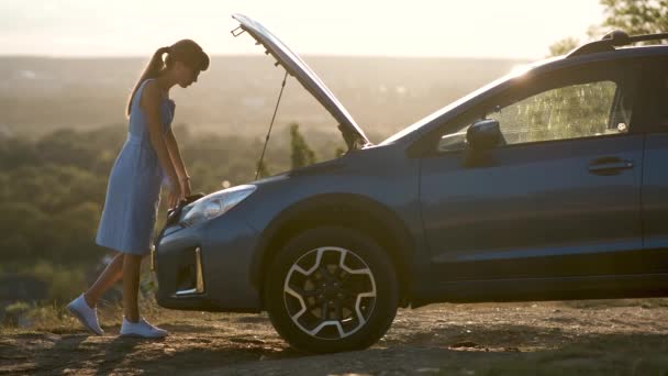 Fiatal női vezető áll egy törött autó mellett nyitott motorháztetővel, akinek problémája van a járművével.. - Felvétel, videó