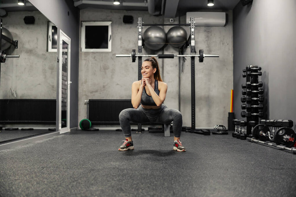 Приседание и сжигание мышц ягодиц и ног. Портрет горячей женщины в спортивной одежде и хорошей физической форме, делающей приседания в изолированном крытом спортзале. Сила и мотивация, фитнес-цель - Фото, изображение