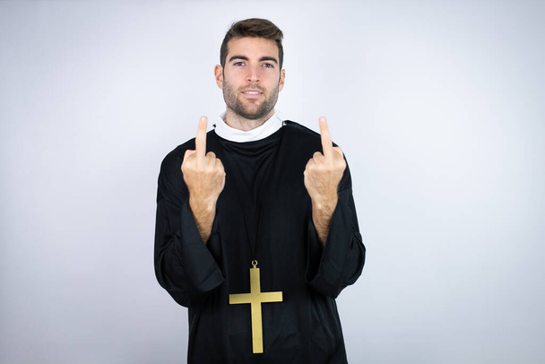 Young hispanic man wearing priest uniform standing over white background showing middle finger doing fuck you bad expression, provocation and rude attitude. screaming excited - Foto, imagen