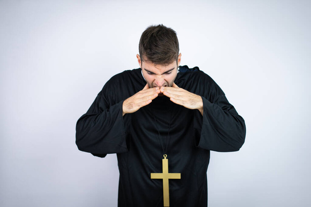Giovane uomo ispanico indossa uniforme sacerdote in piedi su sfondo bianco gridando e urlando forte giù con le mani sulla bocca - Foto, immagini