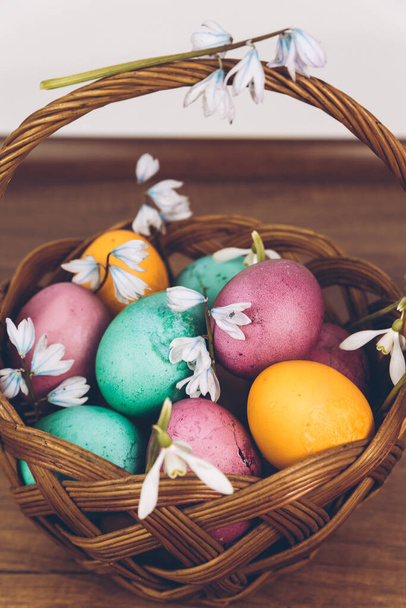 Пасхальный фон. Пасхальные яйца в корзине. Пасхальная корзина с желтыми, зелеными и фиолетовыми яйцами, украшенная подснежниками, стоит на деревянном столе. - Фото, изображение