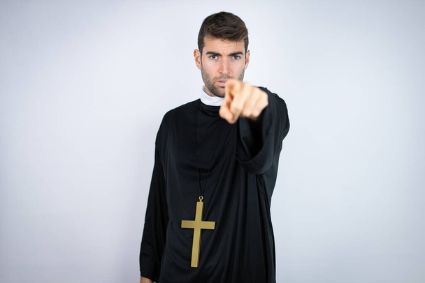 Joven hombre hispano vistiendo uniforme de sacerdote de pie sobre fondo blanco señalando con el dedo a la cámara y a ti, gesto confiado que parece serio - Foto, imagen