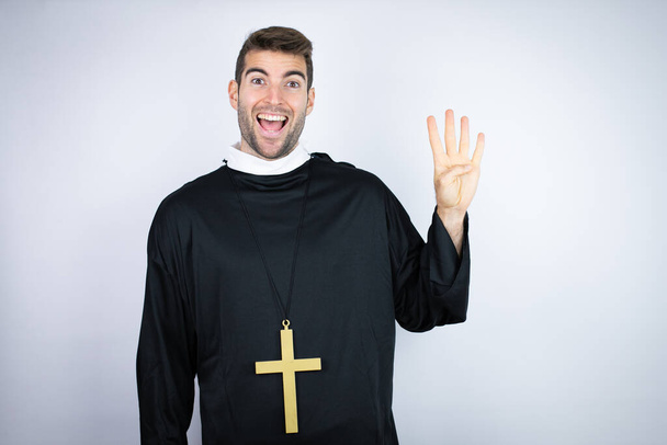 Молодой латиноамериканец в форме священника стоит на белом фоне и показывает пальцами номер четыре, улыбаясь уверенно и счастливо - Фото, изображение