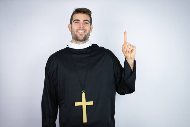 Giovane uomo ispanico indossa uniforme prete in piedi su sfondo bianco mostrando e indicando con le dita numero uno, mentre sorridendo fiducioso e felice - Foto, immagini