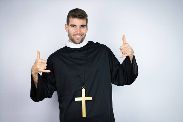 Joven hombre hispano vistiendo uniforme de sacerdote de pie sobre fondo blanco gritando con expresión loca haciendo símbolo de roca con las manos arriba - Foto, Imagen