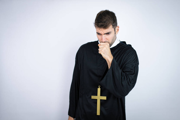 Joven hombre hispano vistiendo uniforme de sacerdote de pie sobre fondo blanco con la mano en la boca porque está tosiendo - Foto, imagen