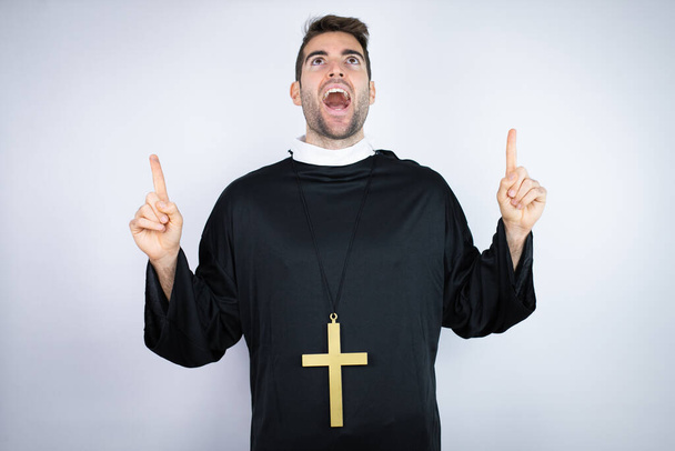Fiatal spanyol férfi, papi egyenruhát visel, fehér háttér felett áll, csodálkozva és meglepve néz a kamerába, és ujjal mutogat felfelé és felemelt karokkal. - Fotó, kép