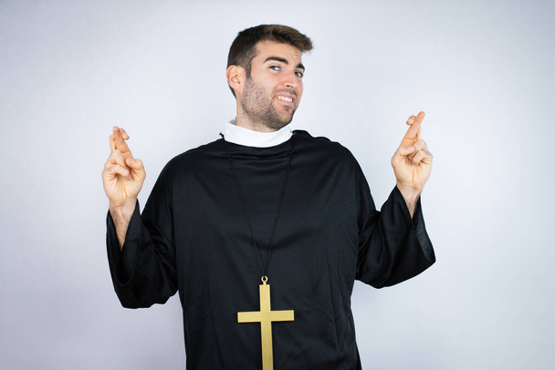 Joven hombre hispano vistiendo uniforme de sacerdote de pie sobre fondo blanco gesto dedo cruzado sonriendo con esperanza y mirando al lado - Foto, imagen