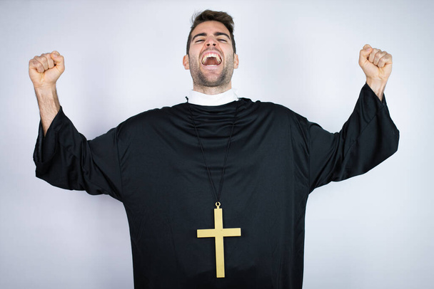 Joven hombre hispano vistiendo uniforme de sacerdote de pie sobre fondo blanco muy feliz y emocionado haciendo gesto ganador con los brazos levantados, sonriendo y gritando por el éxito. - Foto, imagen