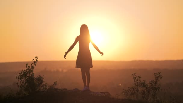 Σκούρα σιλουέτα μιας νεαρής γυναίκας που στέκεται με σηκωμένα τα χέρια πάνω σε μια πέτρα απολαμβάνοντας τη θέα στο ηλιοβασίλεμα το καλοκαίρι. - Πλάνα, βίντεο