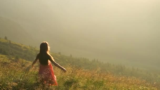 Nuori nainen punaisessa mekossa seisoo nurmettuneella pellolla tuulisena iltana syksyllä vuoret nostaen kätensä nauttien näkymästä luontoon. - Materiaali, video
