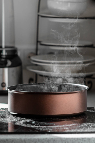 Boiling water with a steam in a brown stewing pan on an electric stove in the kitchen. Blurred background, selective focus. - Photo, Image