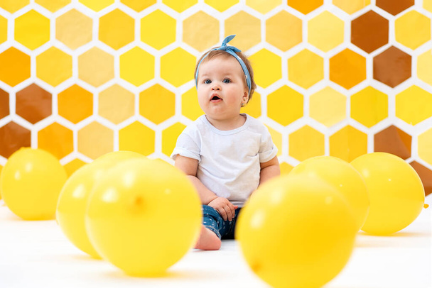 The concept of the World Children's Day. A cute toddler girl is sitting on a floor with yellow balloons. In the background is yellow honeycombs. - Photo, Image