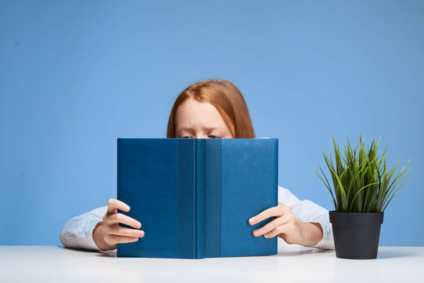 κοκκινομάλλα κορίτσι κάθεται σε ένα τραπέζι με ένα βιβλίο στα χέρια της μαθαίνοντας μαθήματα - Φωτογραφία, εικόνα