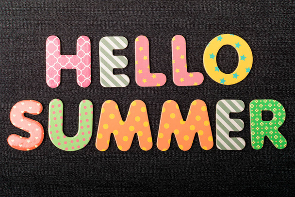 Scheda con Hello Summer parole fatte da lettere di legno colorate vivaci miste su un materiale tessile nero scuro testurizzato che può essere utilizzato come messaggio - Foto, immagini