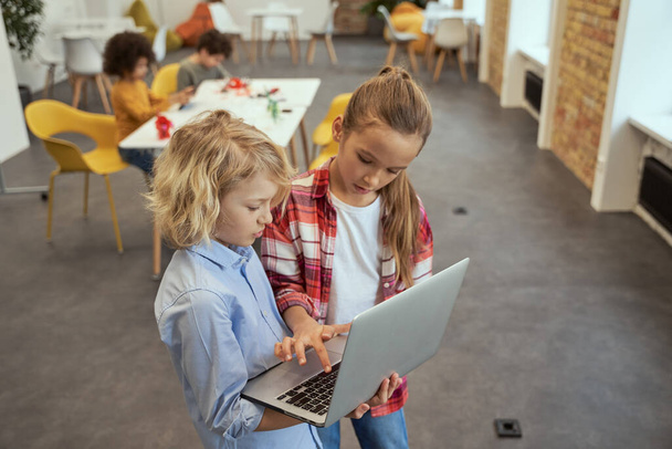 High angle view of two lovely kids, little boy and girl learning, holding laptop and standing in a classroom during STEM lesson - Photo, Image