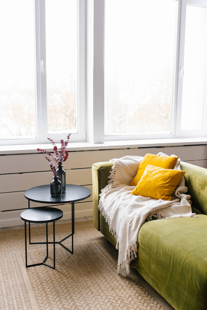 Canapé vert avec oreillers jaunes et couverture beige, table basse avec vase et branches d'eucalyptus. Salon moderne avec grande fenêtre - Photo, image