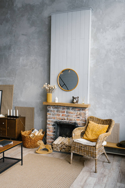 Skandynawski salon w szaro-złotych kolorach z 2021 roku. Wnętrze wiejskiego domu z kominkiem i krzesłem wiklinowym - Zdjęcie, obraz