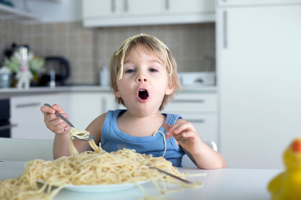 Little blond boy, toddler child, eating spaghetti for lunch and making a mess at home in kitchen - Foto, afbeelding