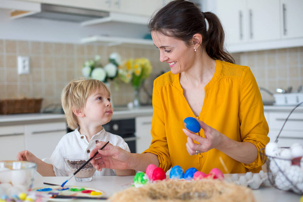 Beautiful blond child, toddler boy, painting easter eggs with mother at home, making easter wreath with rainbow colors - 写真・画像