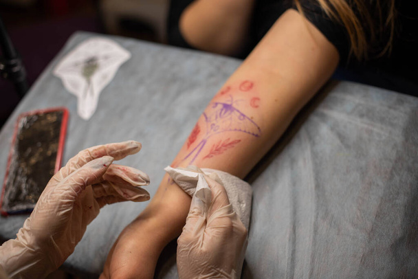 Tatuaż Master rysuje czerwoną farbę na tatuażu klientów. Tatuażysta trzymający różowy tatuaż w czarnych jałowych rękawiczkach i pracujący na profesjonalnej niebieskiej macie. - Zdjęcie, obraz